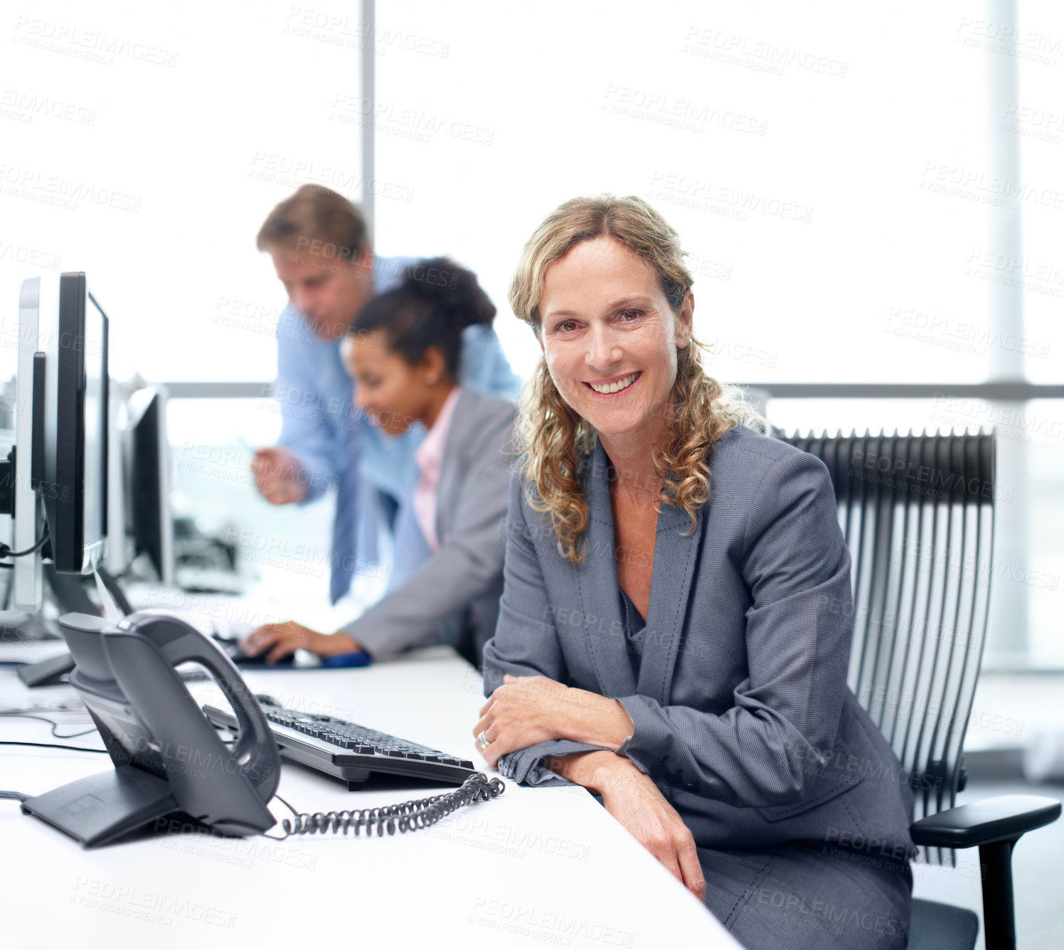 Buy stock photo Positive businesswoman sitting at her desk with her colleagues working in the background - portrait 