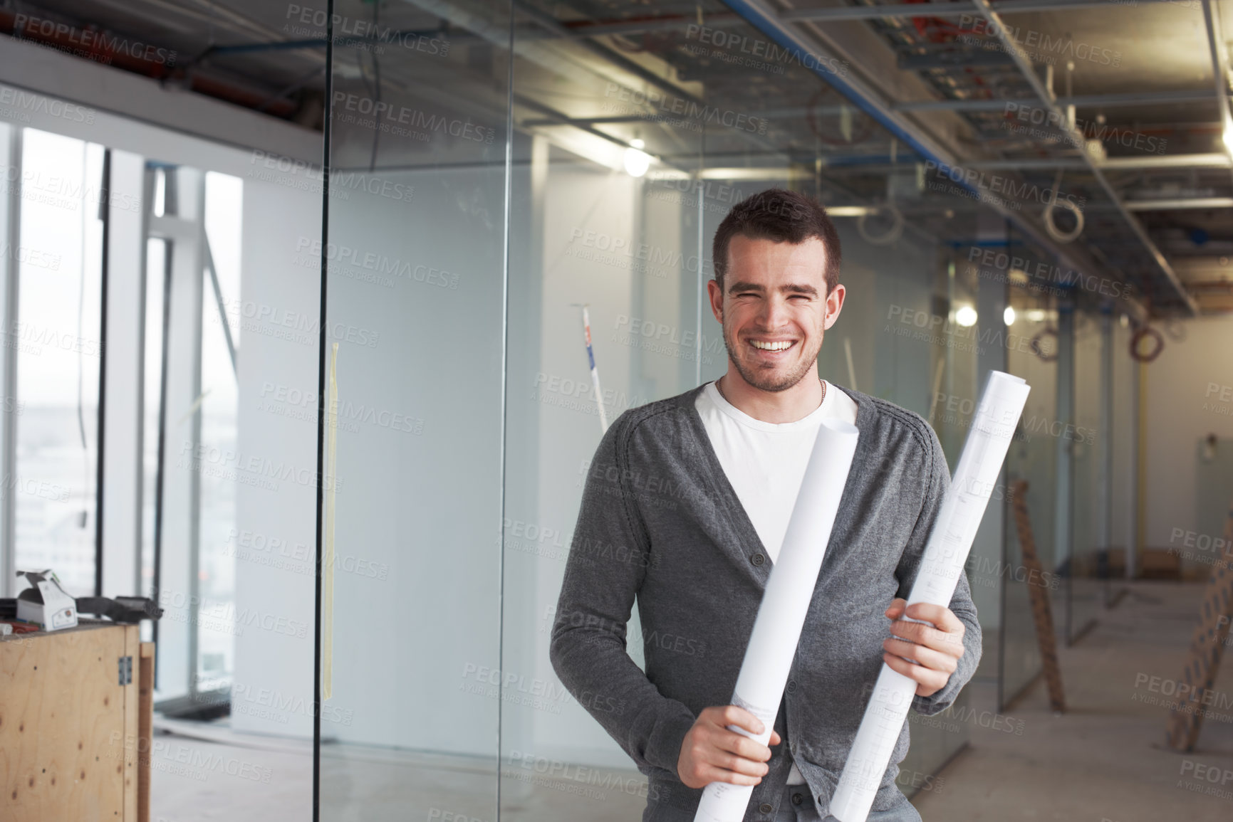 Buy stock photo Young architect standing in a building and holding his plans with a smile