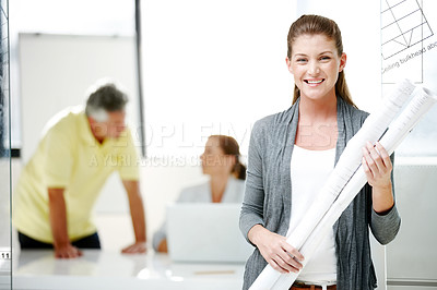 Buy stock photo A young architect standing and holding blueprints while her colleagues work in the background