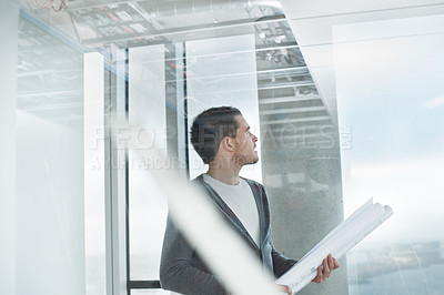 Buy stock photo Young architect standing in a building and holding his plans while looking out the window