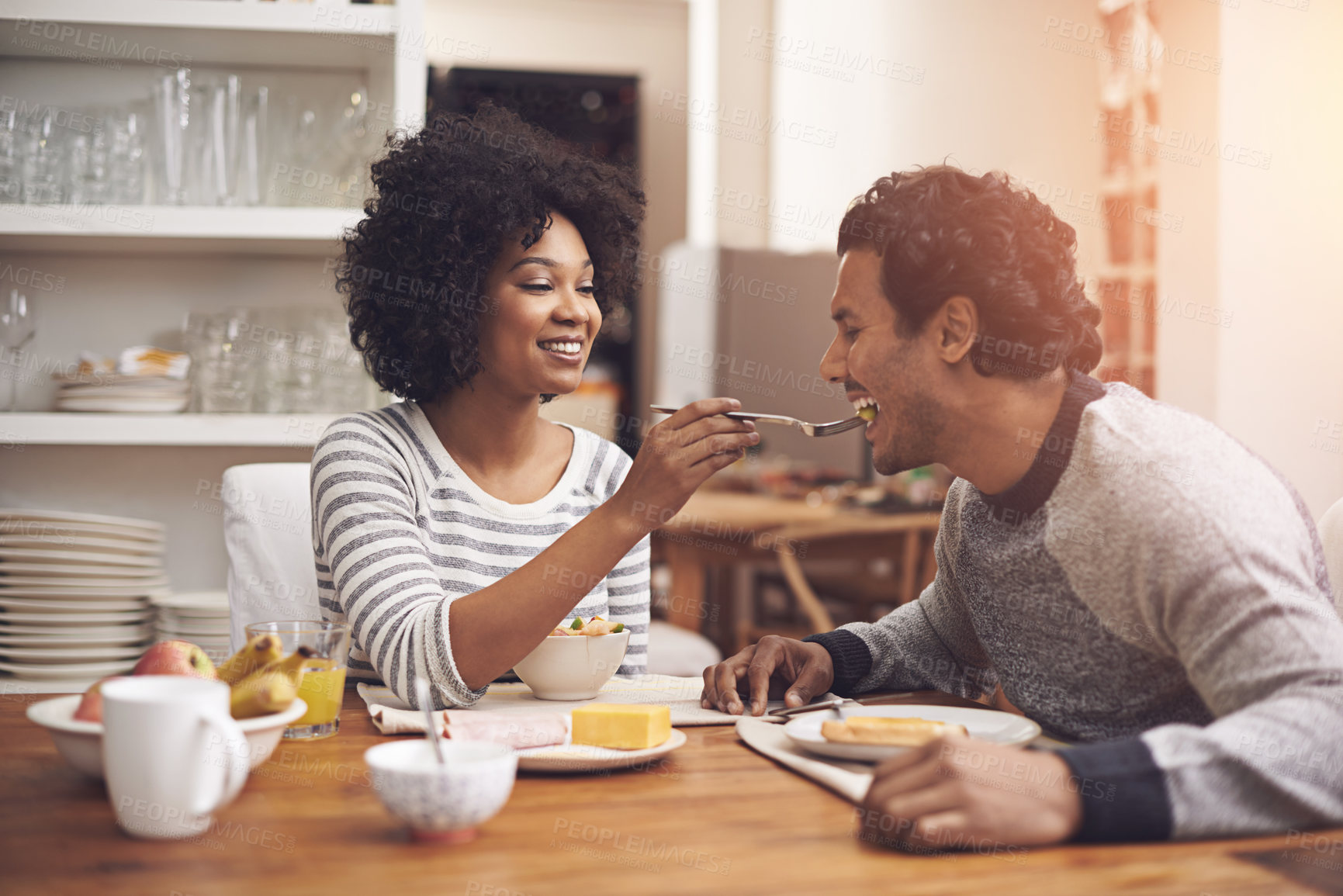 Buy stock photo Breakfast, kitchen and woman feeding husband food for health, wellness and diet at home. Happy, love and young couple bonding and eating meal together on weekend morning at modern apartment.