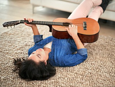 Buy stock photo Shot of a young woman playing the guitar while lying on her living room floor