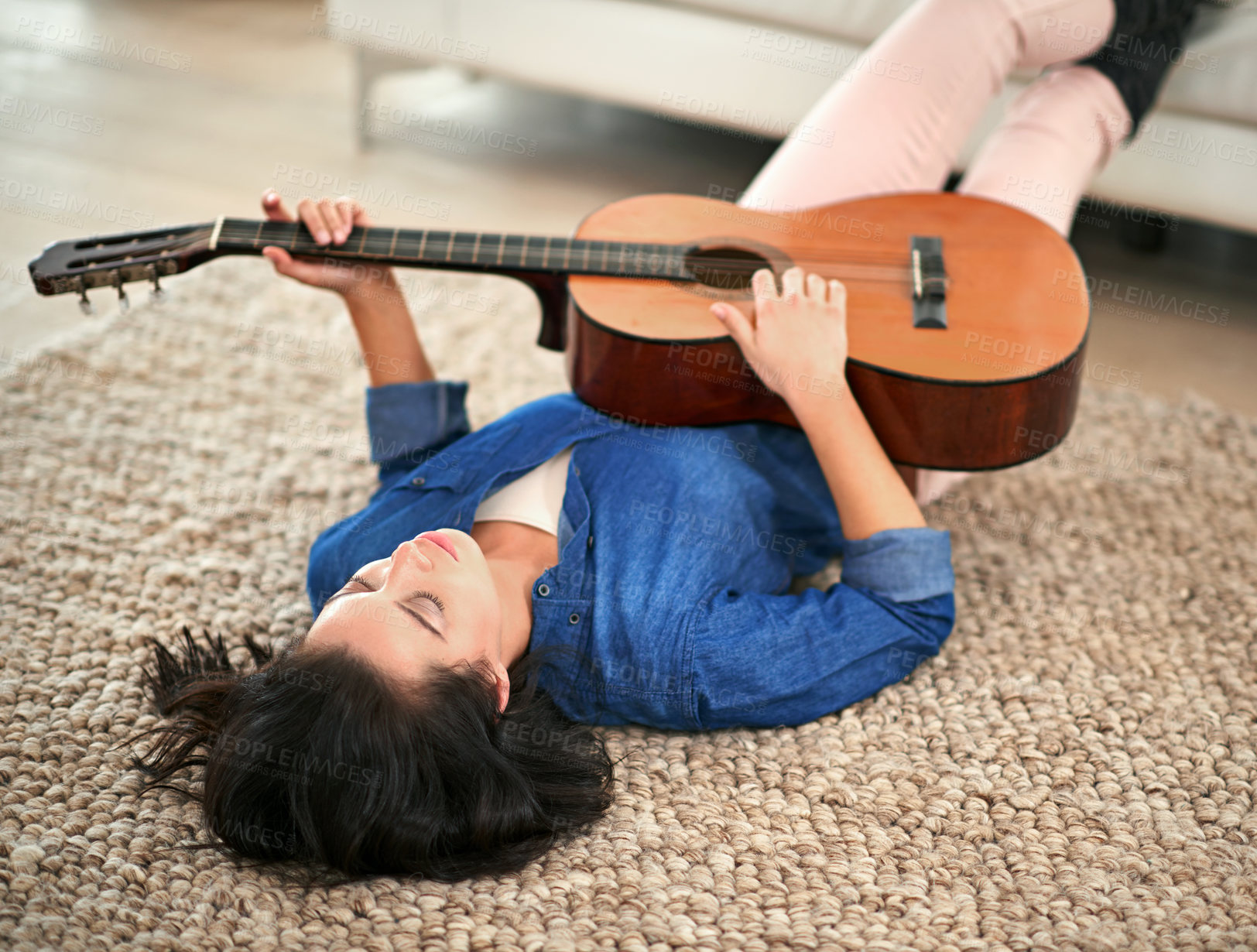 Buy stock photo Shot of a young woman playing the guitar while lying on her living room floor