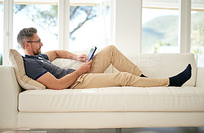 Buy stock photo Shot of a man using his tablet while relaxing on the sofa at home