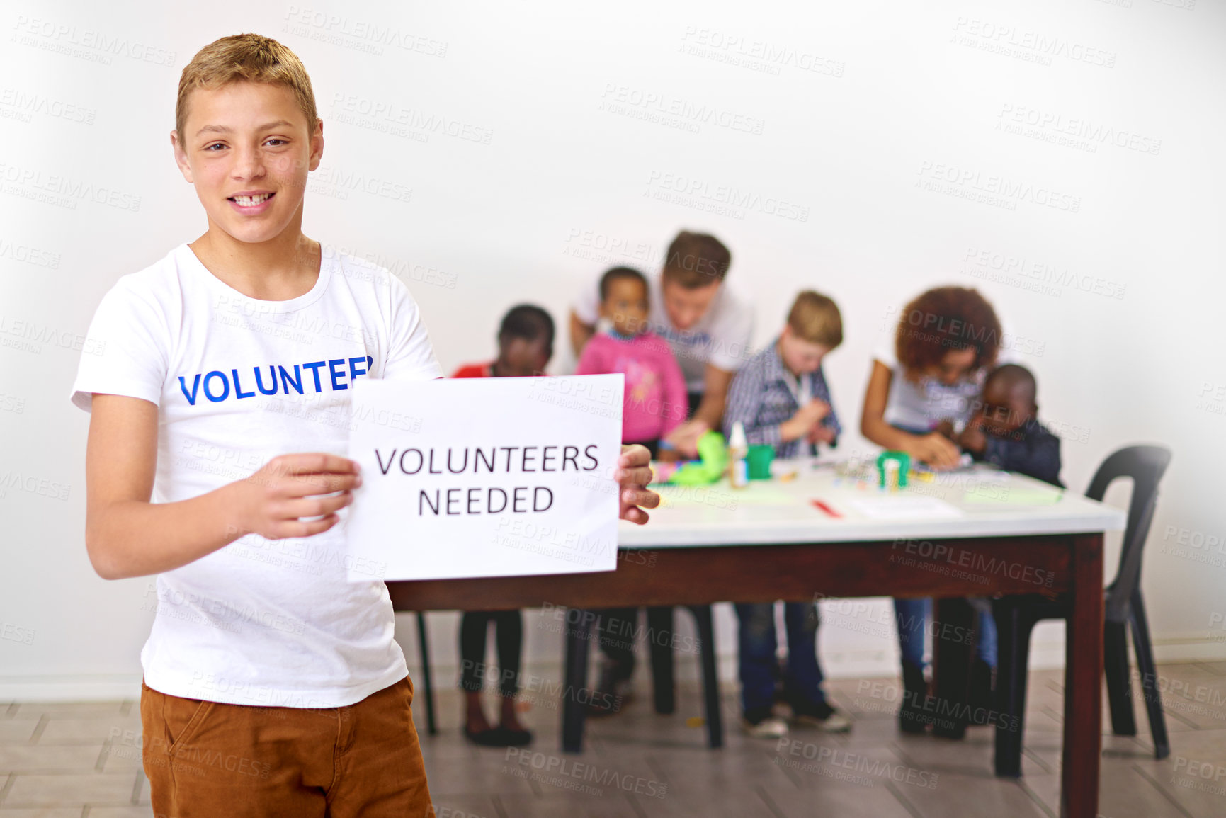 Buy stock photo Portrait of a volunteer holding a 'volunteers needed' sign  with volunteers working with little children in the background