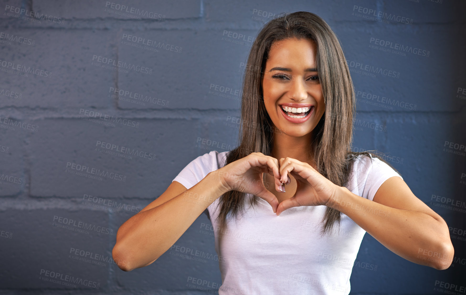 Buy stock photo Portrait of a young woman making a heart gesture with her hands against a brick wall background