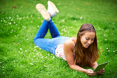Buy stock photo Shot of a young woman using her digital tablet while lying on the grass at the park