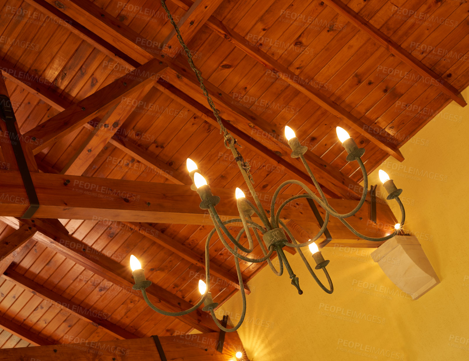 Buy stock photo Below view of an old fashioned chandelier with candle lightbulbs hanging from sturdy wooden roof ceiling inside. Architecture beam detail, interior design, brass, antique light fixture in empty room