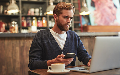Buy stock photo Shot of a young man using a laptop and phone in a cafe