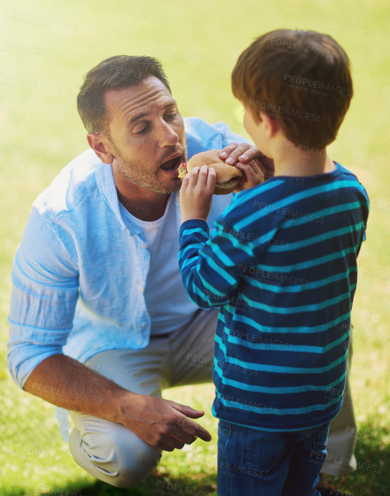 Buy stock photo Grass, man and child with feeding of burger for nutrition, generosity or unhealthy diet together. Family love, father and son with fast food in park for meal sharing, kindness or eating lunch on lawn