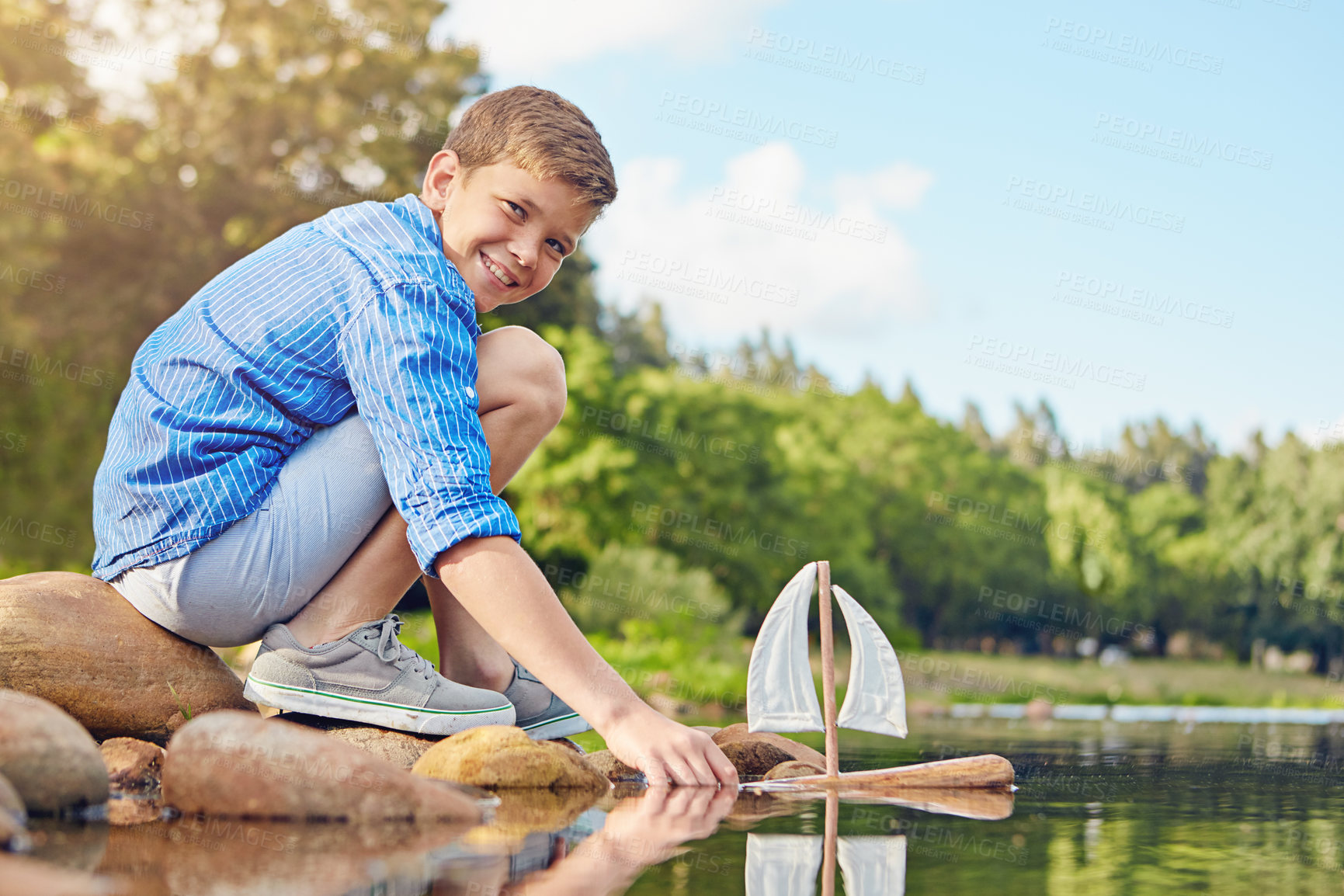 Buy stock photo Shot of a young boy playing with a toy boat by the water