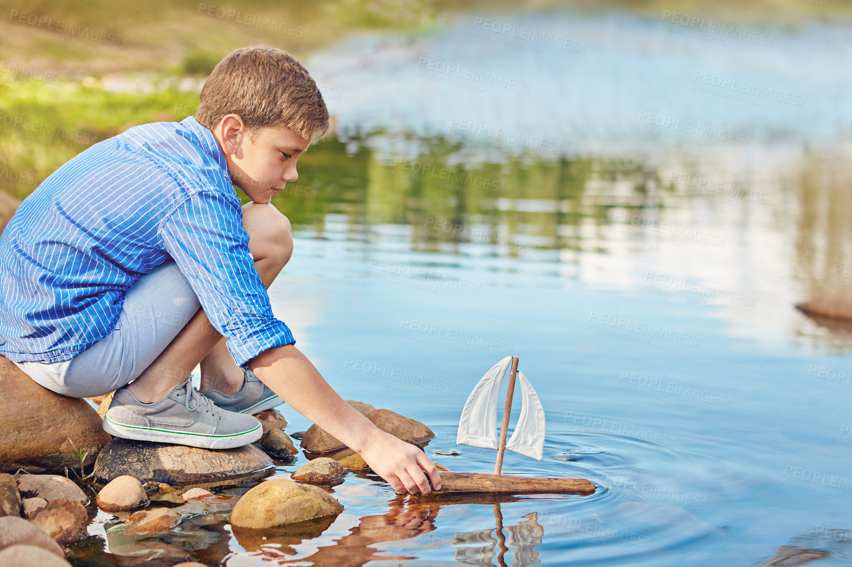 Buy stock photo Shot of a young boy playing with a toy boat by the water