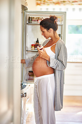 Buy stock photo Shot of a pregnant woman standing in front of a fridge with a cupcake in her hand