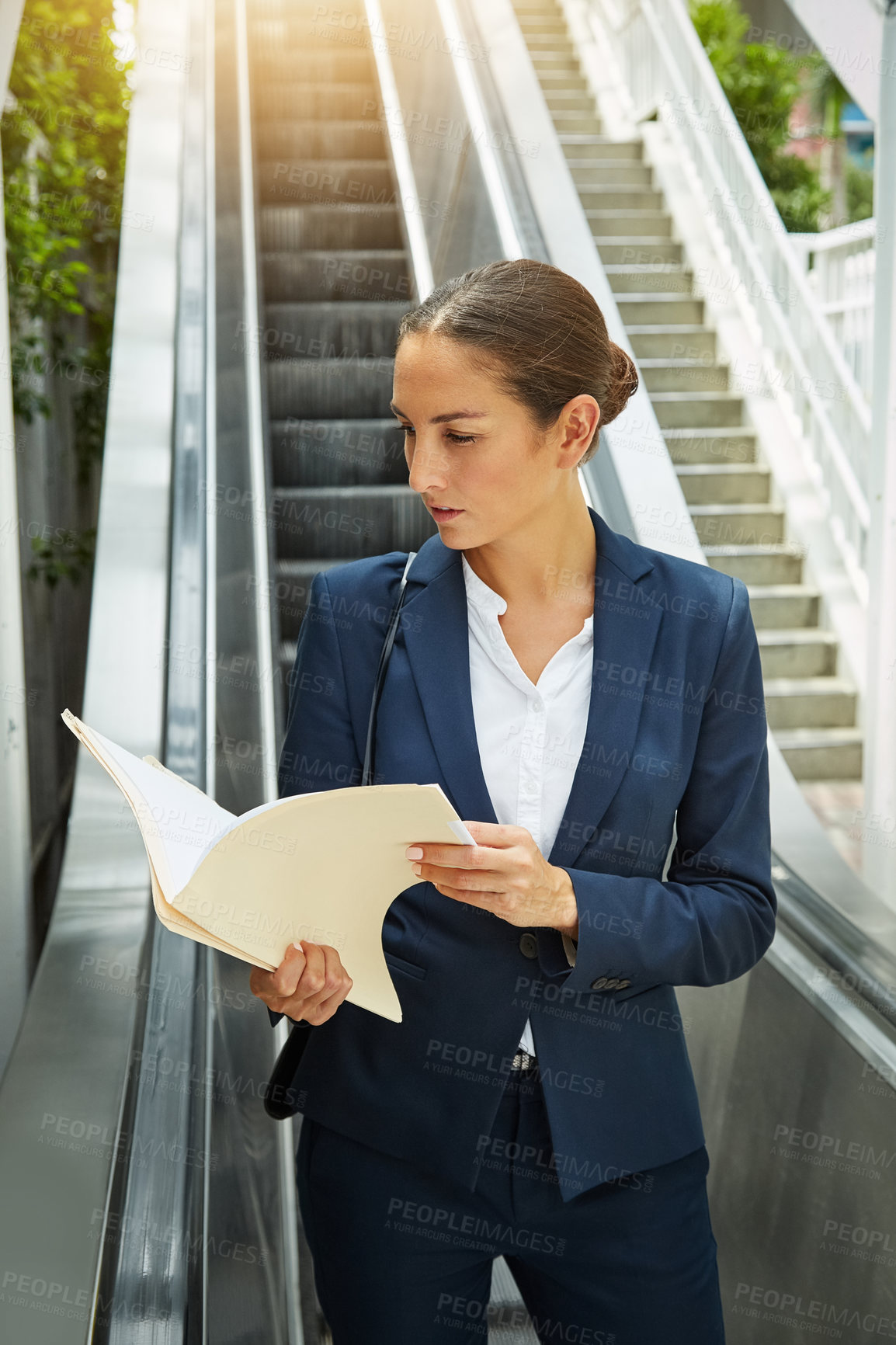 Buy stock photo Shot of a young businesswoman reading paperwork on an escalator