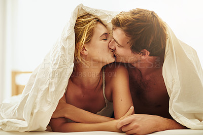 Buy stock photo Shot of a young couple sharing a kiss under the covers in bed