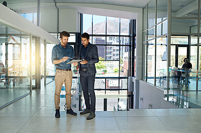 Buy stock photo Shot of two colleagues talking together over a digital tablet while standing in a modern office