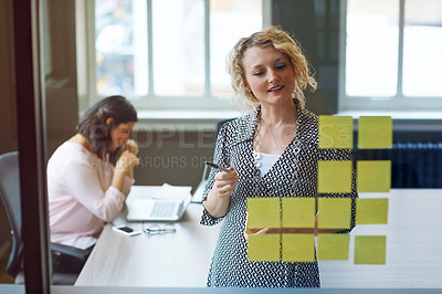 Buy stock photo Shot of a businesswoman reading adhesive notes on a glass wall with a colleague in the background