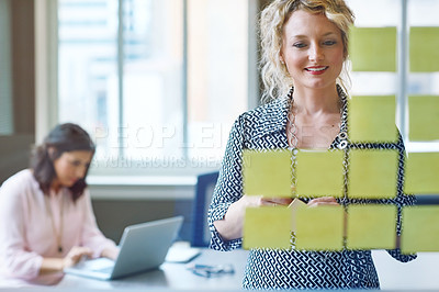Buy stock photo Shot of a businesswoman reading adhesive notes on a glass wall with a colleague in the background