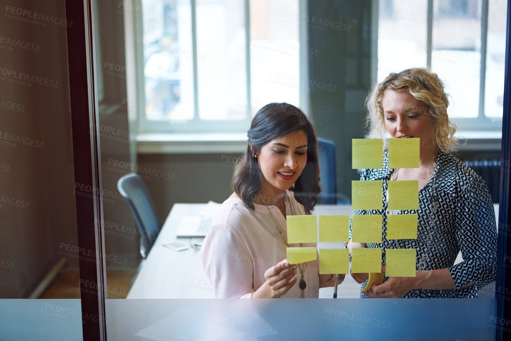 Buy stock photo Shot of two businesswomen brainstorming with adhesive notes on a glass wall in an office