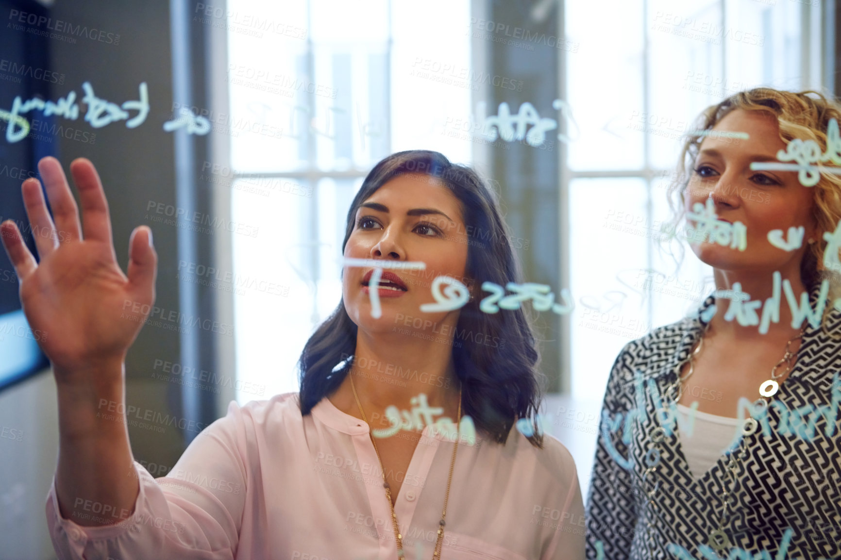 Buy stock photo Shot of two businesswoman brainstorming on a glass wall in an office