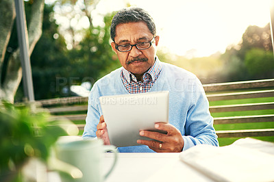 Buy stock photo Shot of an older man using a digital tablet while having his breakfast outdoors