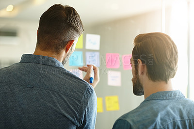 Buy stock photo Rearview shot of two young businessmen brainstorming on a glass wall while standing in an office