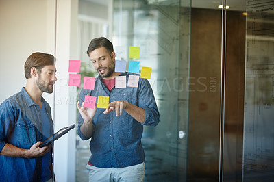 Buy stock photo Shot of two young businessmen brainstorming on a glass wall while standing in an office