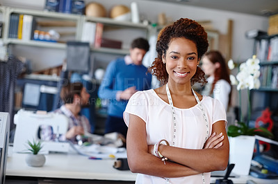 Buy stock photo Portrait of a young fashion designer standing with her arms folded while her colleagues work in the background