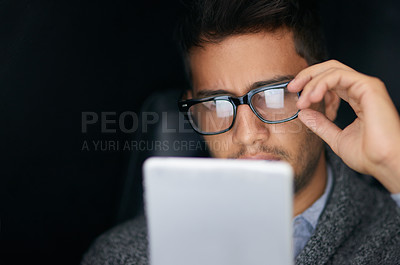 Buy stock photo Shot of a young man wearing glasses using a digital tablet in the dark