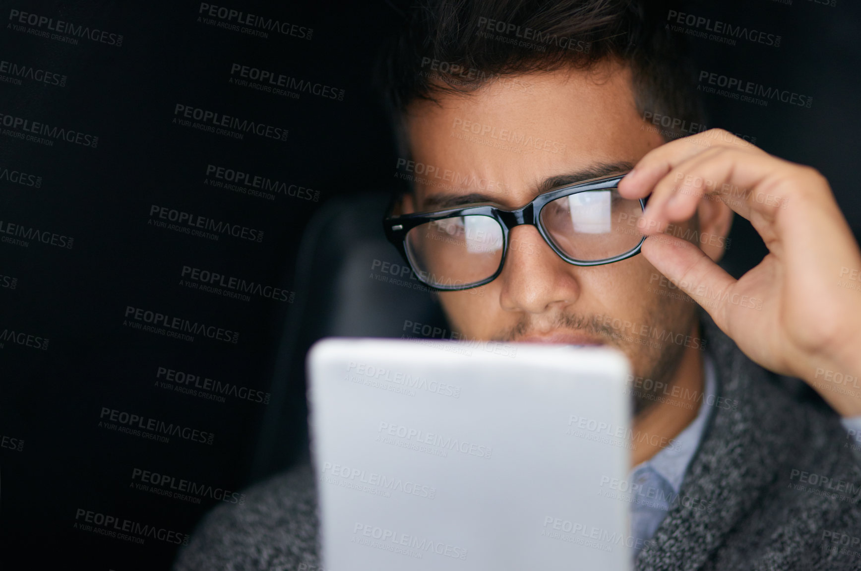Buy stock photo Shot of a young man wearing glasses using a digital tablet in the dark