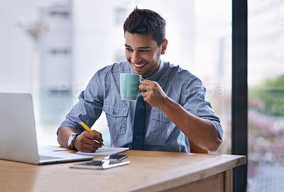Buy stock photo Shot of a young businessman writing notes and drinking coffee while working on a laptop in an office