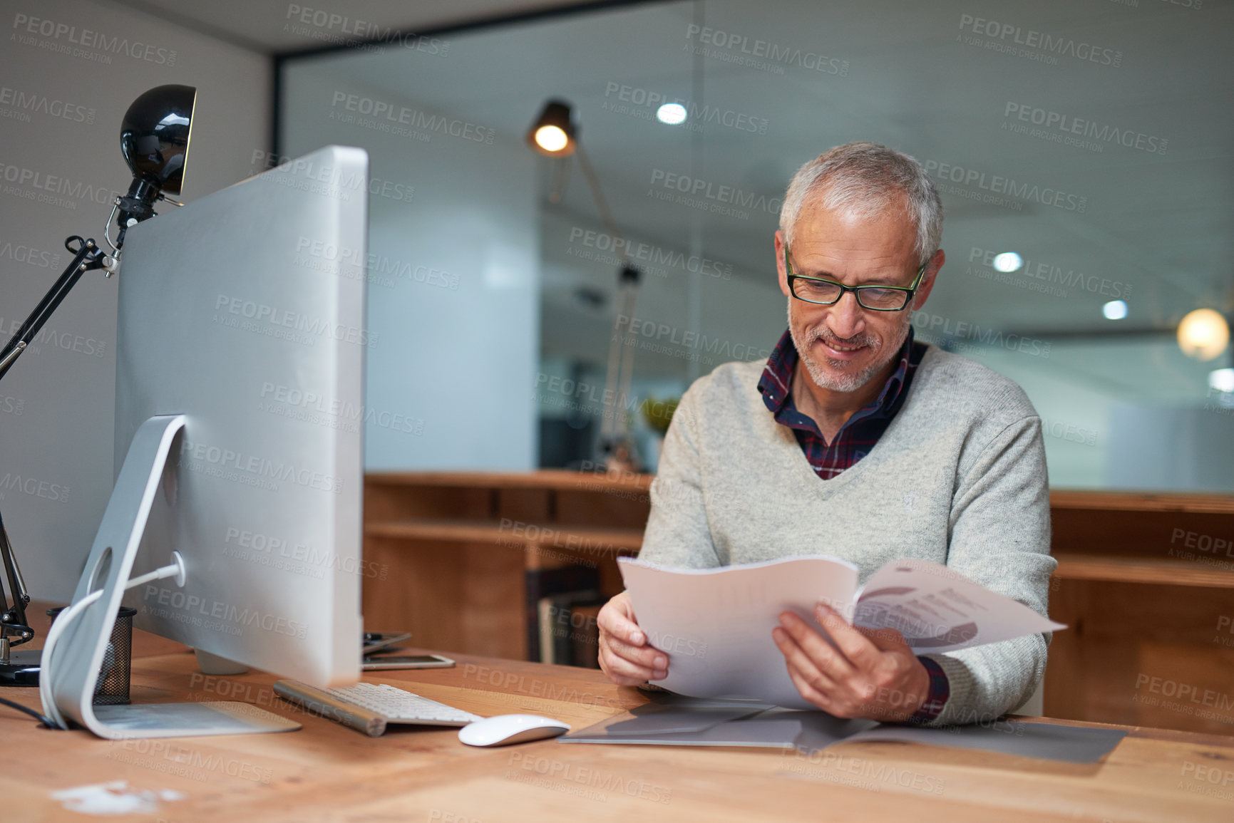 Buy stock photo Shot of a smiling mature businessman reading paperwork while sitting at his desk in an office