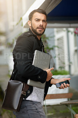 Buy stock photo Shot of a handsome young man carrying his phone and laptop while out in the city