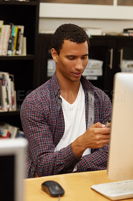 Buy stock photo Cropped shot of a university student texting on a phone while working on a computer in the campus library