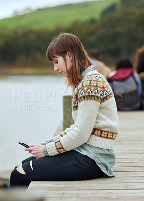 Buy stock photo Shot of a young woman sending a text message while sitting on the pier by a lake