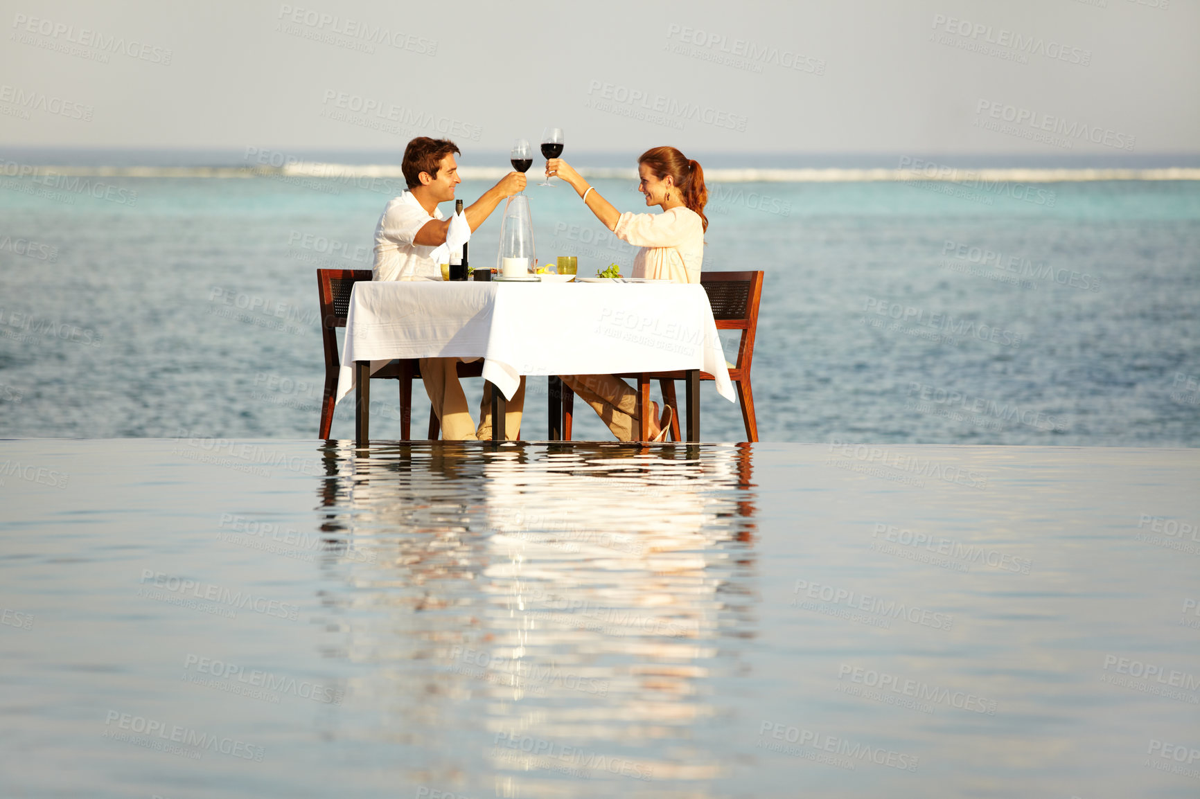 Buy stock photo A happy couple clinking their wine glasses while sitting outside surrounded by water