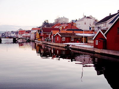 Buy stock photo Small houses in a town near a quiet lake with a bridge in the background. Scandinavian maritime town on a sunny morning. A village on a lake surrounded by a snowy landscape with water and hills