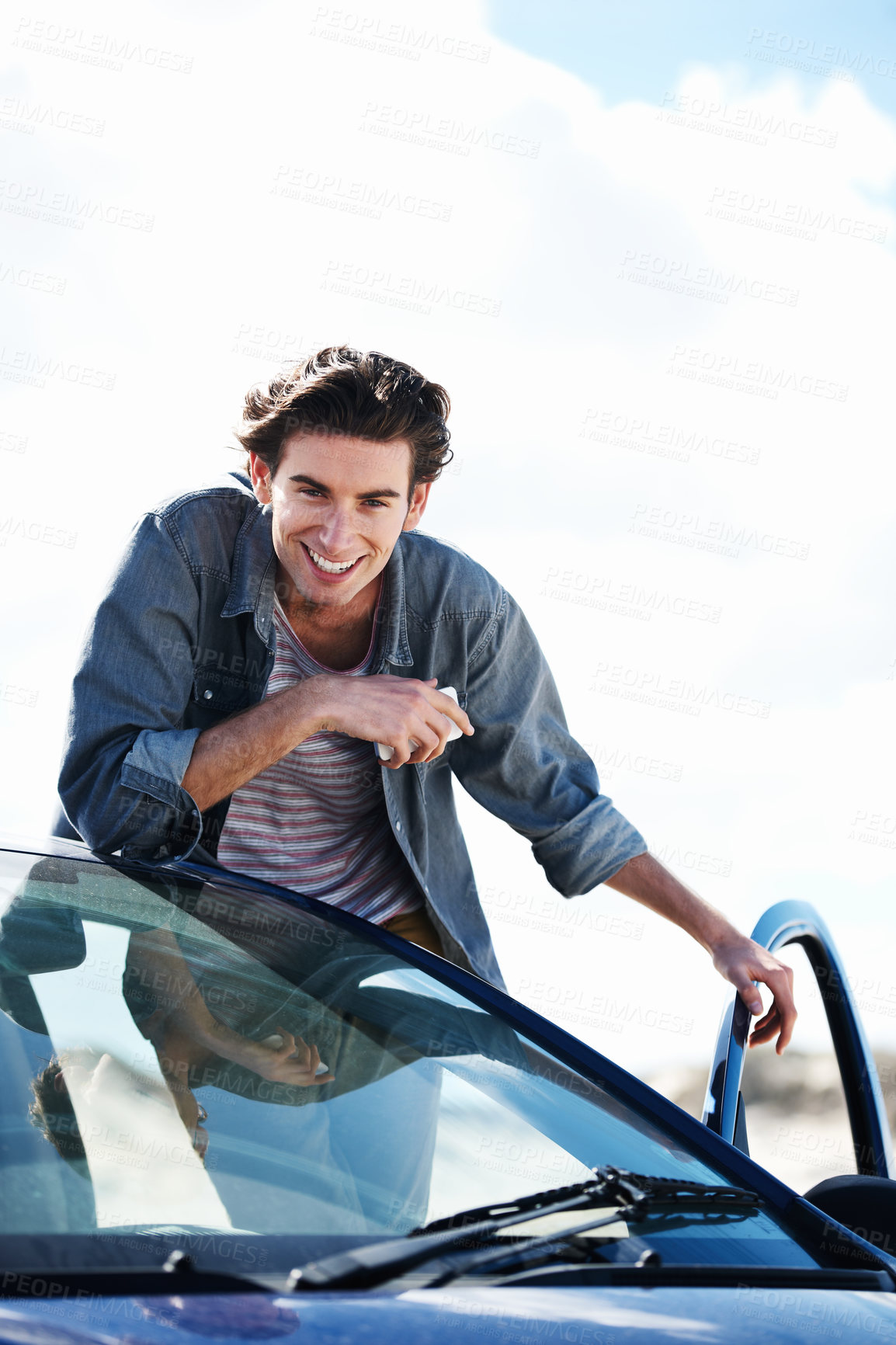 Buy stock photo Happy young man holding a cellphone and leaning on the roof of his car while smiling at the camera