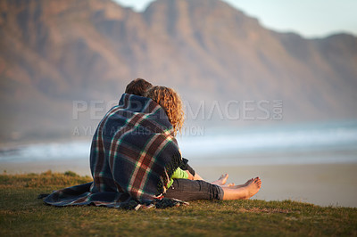 Buy stock photo Cute young couple wrapped in a blanket while sitting on a riverbank