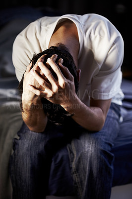 Buy stock photo A young man hanging and holding his head in desparation at the foot of his bed