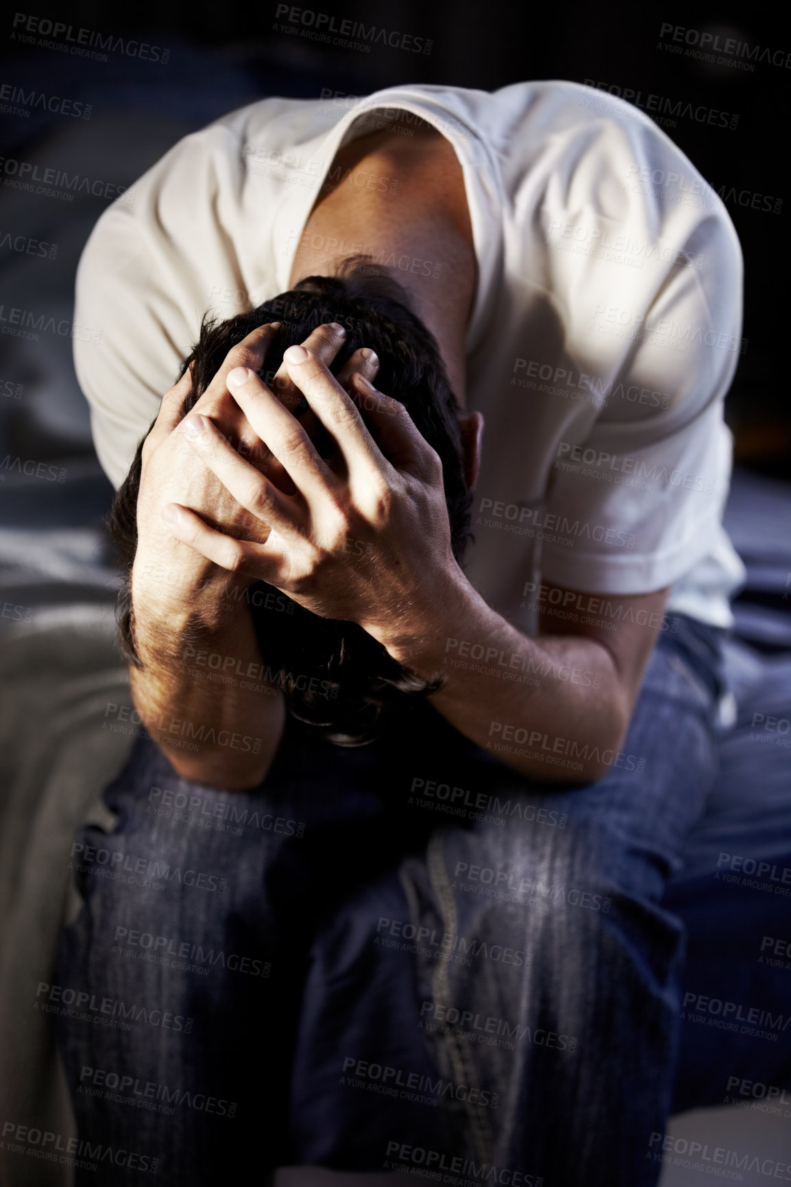 Buy stock photo A young man hanging and holding his head in desparation at the foot of his bed