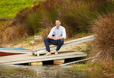 Buy stock photo Portrait of a mature wine maker sitting on a jetty with a glass of red wine