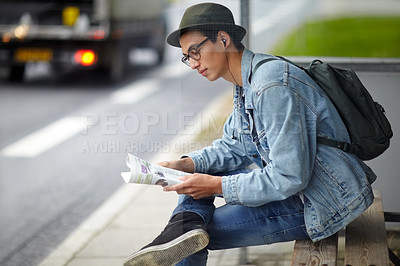 Buy stock photo Hip young guy sitting at a bus stop while looking at a map