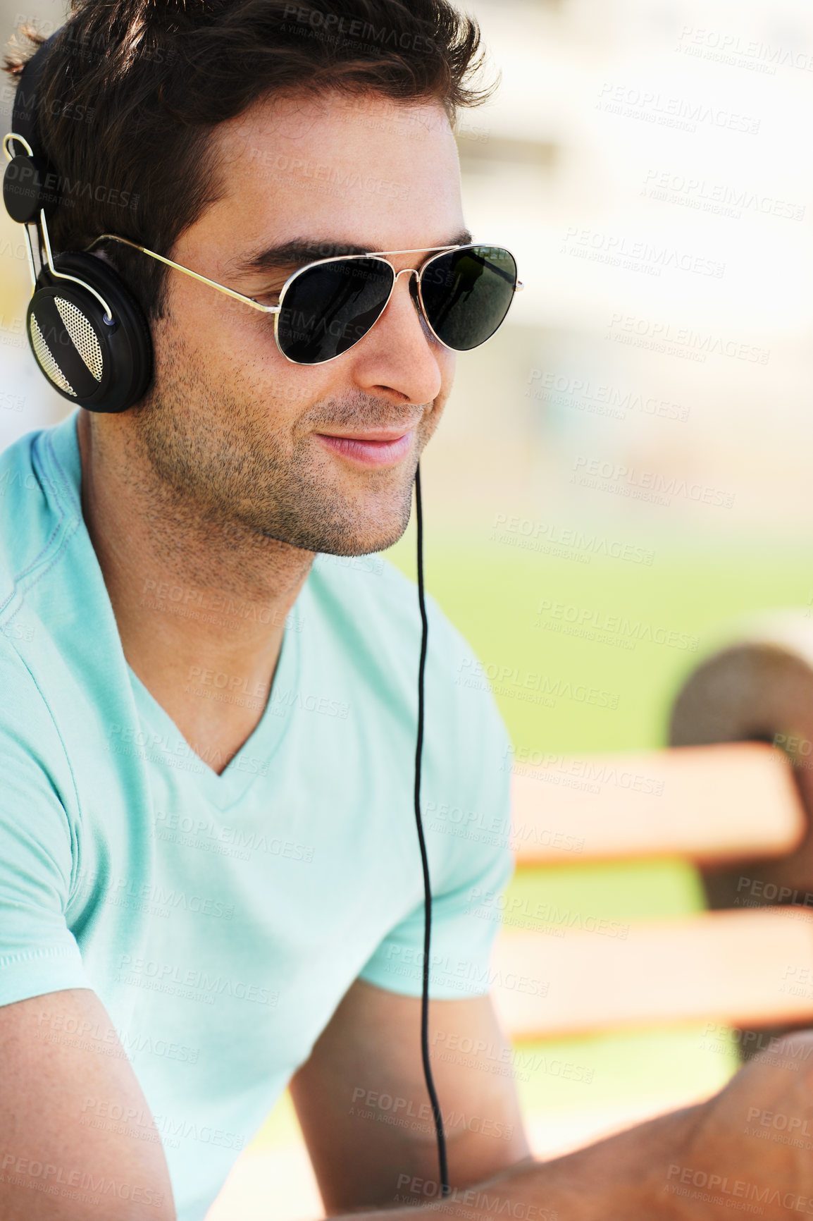 Buy stock photo A smiling young man wearing sunglasses sitting on a bench in a park and listening to music through his headphones