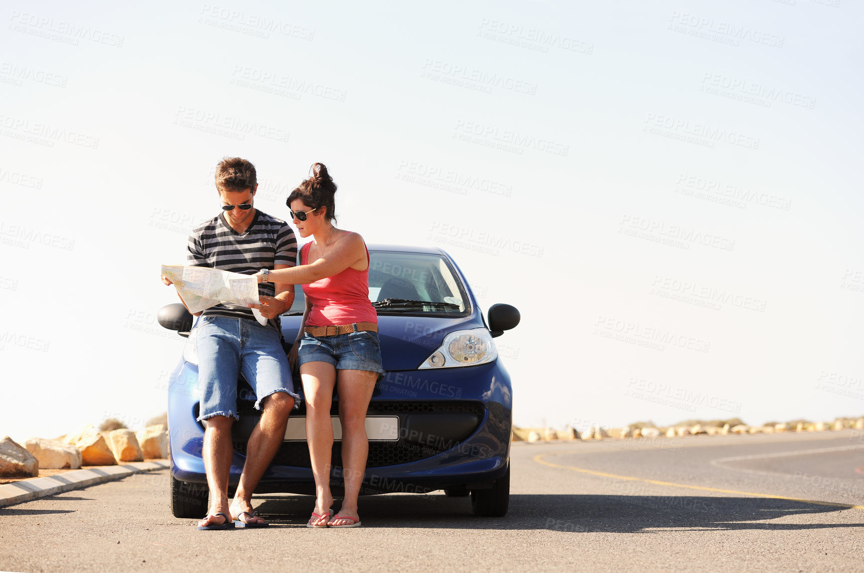 Buy stock photo A man and a woman parked on the side of the road and reading a map together while seated on the bonnet of their car