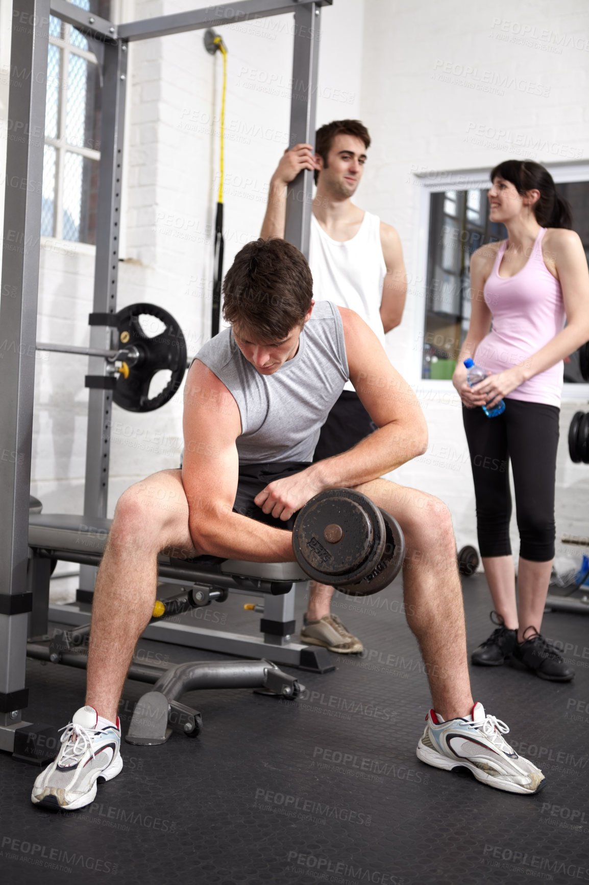 Buy stock photo young bodybuilder lifting a weight while two people chat behind him in the gym