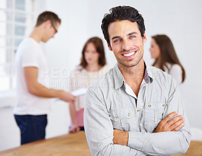 Buy stock photo Portrait of a positive-looking young business professional standing with his arms folded with coworkers talking in the background