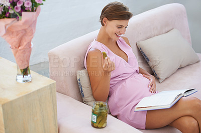 Buy stock photo A happy pregnant woman satisfying a craving for pickles while reading on the sofa