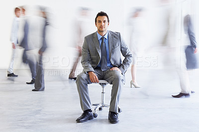 Buy stock photo Conceptual portrait of a young executive sitting as colleagues stream passed him hastily