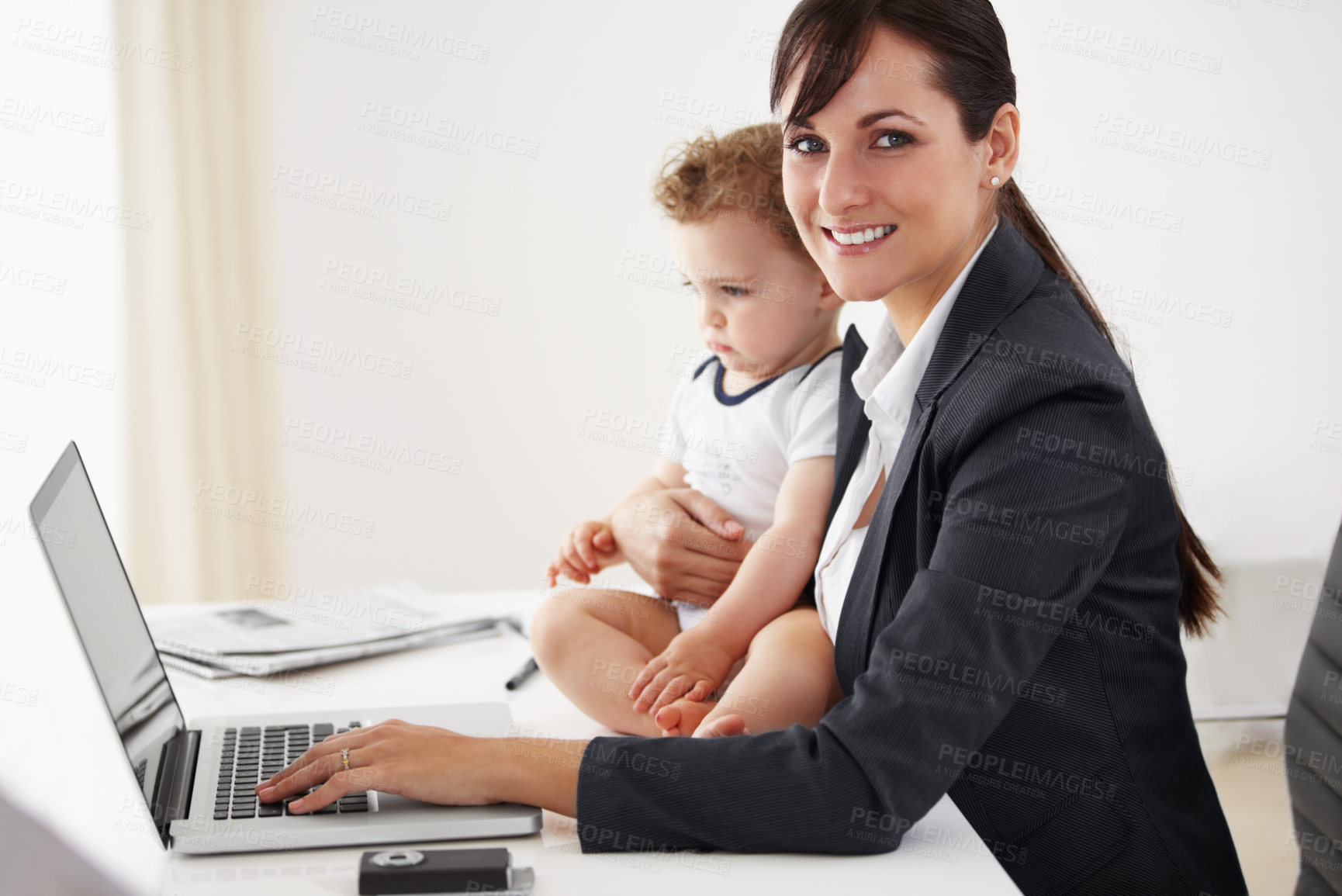 Buy stock photo Young working mom smiling at the camera while holding a camera and typing on her laptop